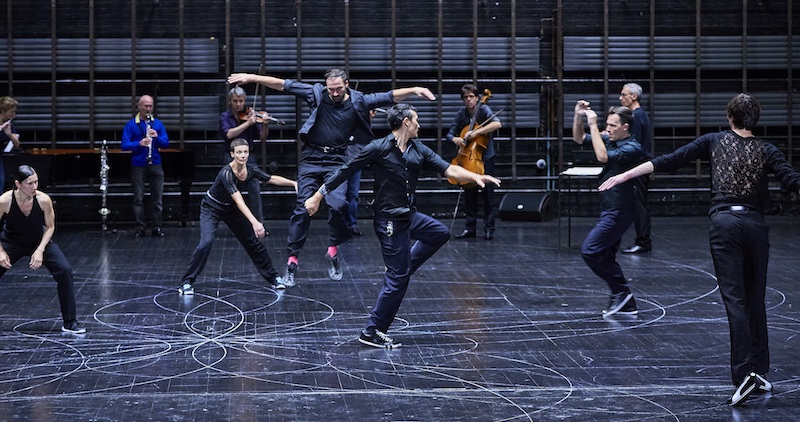 Several dancers in black and sneakers dance different moves while musician play in the background.